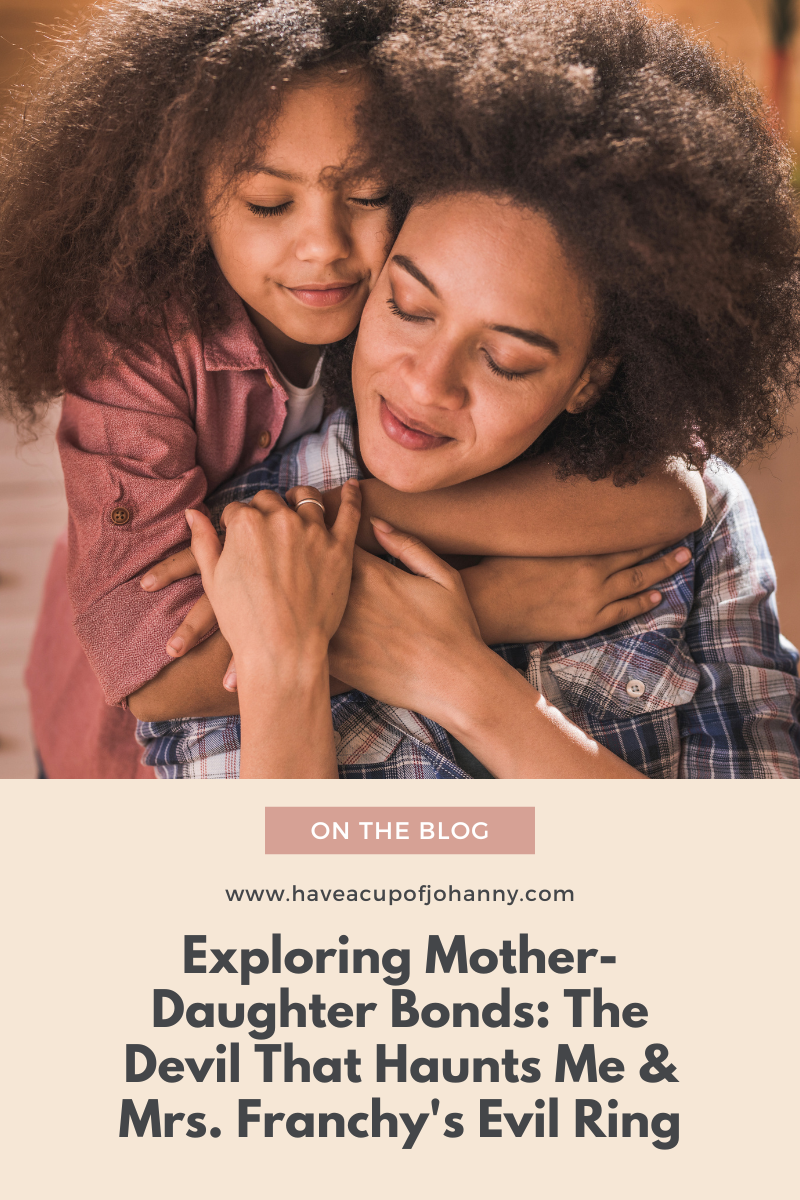 A warm and affectionate moment between a mother and daughter, with the daughter hugging her mother from behind, both smiling contentedly. The image is used as the feature for a blog post titled 'Exploring Mother-Daughter Bonds: The Devil That Haunts Me & Mrs. Franchy’s Evil Ring' on the website haveacupofjohanny.com. The post is highlighted as a new entry on the blog, inviting readers to engage with the content.